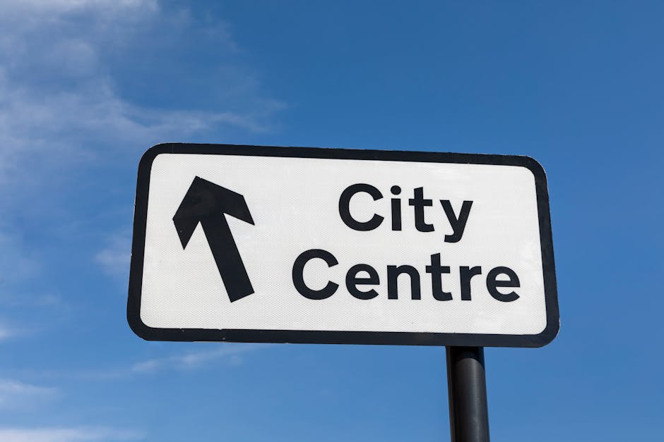 A rectangular white road sign with a black border and black text displaying 'City Centre' and an upward-pointing arrow, mounted on a black pole against a clear blue sky with a few wispy clouds. The sign is positioned outdoors, likely near a main road or highway, indicating the direction to the city centre. The image is relevant to house removals and relocation services provided by Removal Companies Edgware, as it signifies navigation and moving logistics involved in home relocation or furniture transport, with the sign situated in an open environment reflecting the urban setting of a typical move or delivery route.