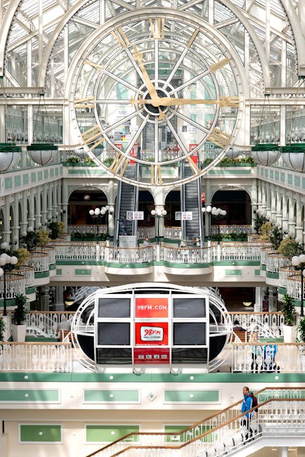 Inside the historic Broadwalk Shopping Centre in Edgware, the image displays a large, ornate clock with gold Roman numerals and clock hands mounted on a glass and metal framework ceiling. The interior features multiple levels with decorative white railings, green panels, and spherical light fixtures. On the ground floor, a temporary office or booth with a red and white 98FM sign is positioned near the staircase. A person wearing a blue jacket and carrying a bag is seen walking along a staircase on the right side. The surroundings include elegant architectural details such as columns and hanging lights, suggesting a setting where home relocation or furniture transport companies, like Removal Companies Edgware, might coordinate moving or packing services within this distinctive, spacious environment.