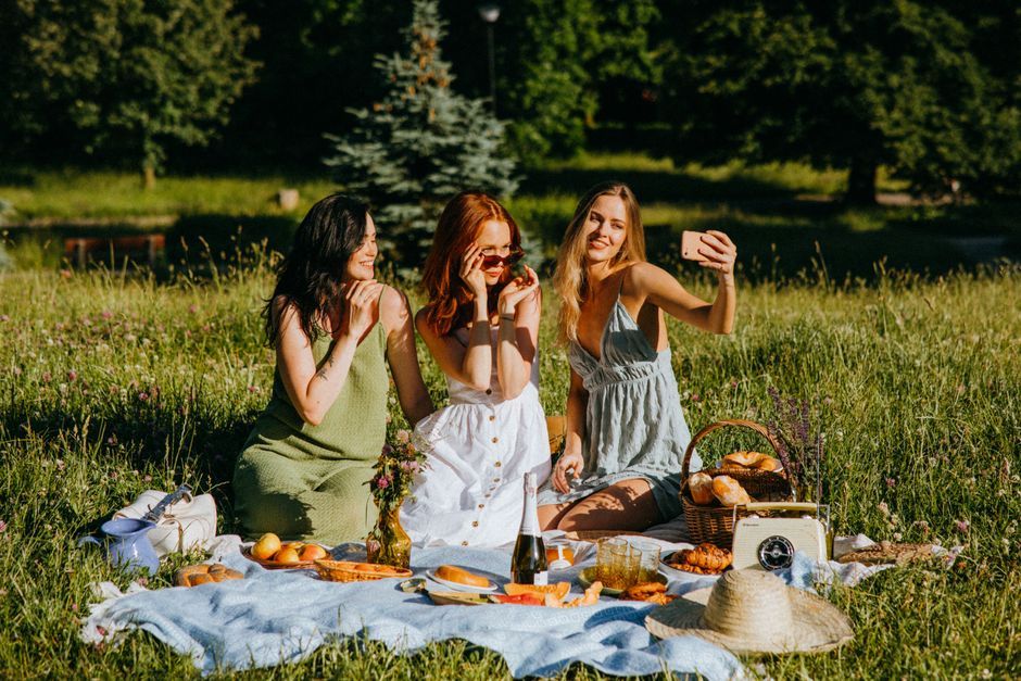 Three young women sit on a blue picnic blanket outdoors in a grassy park area with tall trees and a small pine tree in the background during daytime. They are surrounded by a variety of picnic items, including a wicker basket filled with bread rolls, a glass bottle of drink, a vintage camera, a wide-brimmed straw hat, and plates with sandwiches and pastries. One woman with long blonde hair wears a light gray sleeveless dress and is taking a selfie with her smartphone, while another with long dark hair in a green dress and a third with red hair in white clothing enjoy the moment, adjusting sunglasses and smiling. The scene is well-lit with natural sunlight, capturing a casual, relaxed atmosphere suitable for illustrating a moving or packing-related theme in the context of house relocations or outdoor gatherings supported by Removal Companies Edgware.