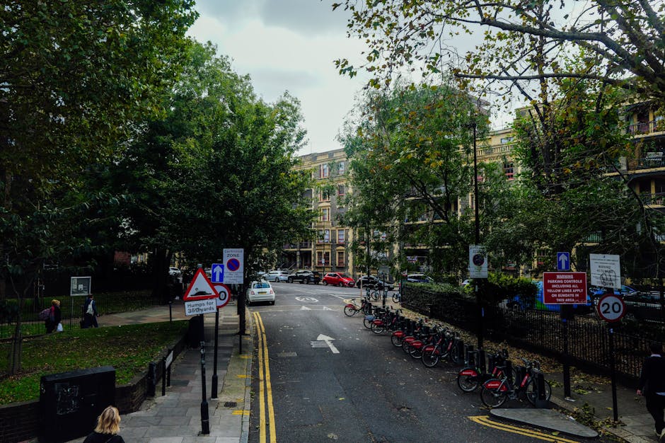 A street scene outside a residential building in Canons Park, Edgware, showing an urban road with parked bicycles secured to metal racks on the sidewalk. Several street signs are visible, including parking restrictions, a no stopping zone, and a 20-mph speed limit, along with directional arrows indicating traffic flow. The road surface appears slightly wet, with yellow lines marking parking restrictions. In the background, there are multi-storey apartment blocks with balconies, surrounded by trees with green and some yellowing leaves, suggesting late summer or early autumn. Pedestrians are walking on the sidewalk, and a few cars are parked or driving along the street. The image reflects an everyday urban environment often associated with house removals or furniture transport, relevant to services like those offered by Removal Companies Edgware.