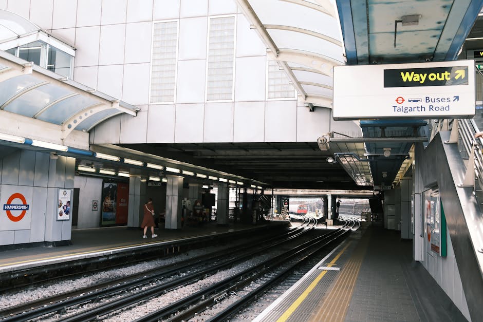 Exterior view of Edgware tube station showing the entrance and train tracks, with a pedestrian walking on the platform, area signage including a 'Way out' sign and a direction sign for buses towards Talgarth Road, and parts of the station's canopy overhead. The scene captures the typical features of a London underground station, with tiled walls, station lighting, and structural elements designed for passenger flow. This setting reflects an urban environment suitable for home relocation services provided by Removal Companies Edgware, highlighting the logistics involved in moving furniture and household items via transport connections near the station, especially during a moving day.