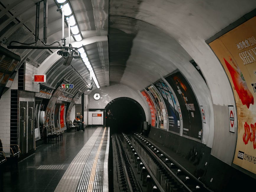 Inside the underground station platform near Edgware tube station, the curved tunnel walls are tiled with white and black tiles, illuminated by overhead fluorescent lights. Along the left side, there are metal benches with a few passengers seated, some wearing high-visibility vests. To the right, colorful advertisements are displayed on the tunnel walls, including one prominently featuring red and yellow hues. The platform surface is made of dark material, with tactile paving strips near the edge for safety. In the background, the dark tunnel opening leads to the underground rail tunnel. Visible equipment includes a small clock hanging from the ceiling, informational signs, and a set of stairs or escalator at the far end. This scene captures the typical environment where house removals might involve the loading and transportation of furniture and boxes, with staff possibly preparing to load items onto a train or transit vehicle as part of a home relocation process. Removal Companies Edgware would coordinate such logistics to ensure secure and efficient packing and furniture transport through this station environment.
