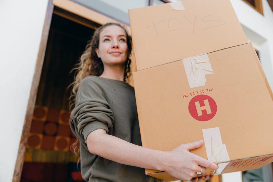 A young woman with long, curly hair and a neutral expression is inside a home, carrying three cardboard boxes stacked one on top of the other during a home relocation. The top box has the word 'toys' handwritten on it in pencil, while the middle and bottom boxes are sealed with packing tape and feature printed markings, including a red circle with an 'H' inside. The woman is wearing a casual, long-sleeved grey top with the sleeves rolled up and is holding the boxes securely with both hands, positioned near a doorway with a wooden frame. Inside, the room has a painted wall, and the background includes a glimpse of a decorated wall with circular patterns. The lighting appears natural, highlighting her focused expression as part of the packing and moving process, exemplified by furniture and packing materials typical for house removals, demonstrating careful handling of packing and transport tasks associated with local house removals in Edgware.